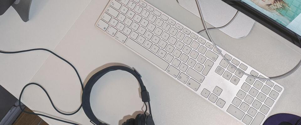 Computador visto de cima, com teclado, fone de ouvido e monitor em uma mesa. 