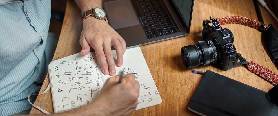 Diretor de arte desenhando fontes em caderno. Na mesa, vemos uma câmera e um notebook. 