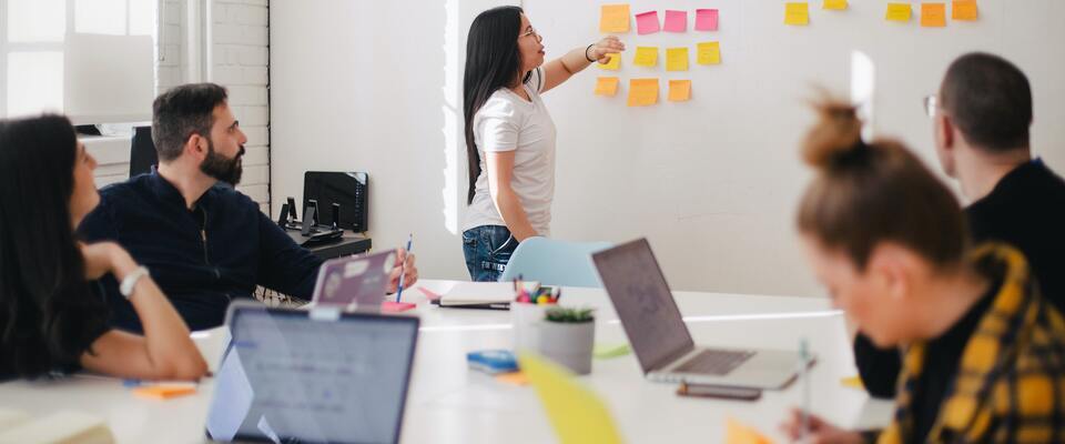 Equipe reunida em sala de reuniões, enquanto uma mulher interage com post its pregados na parede. 