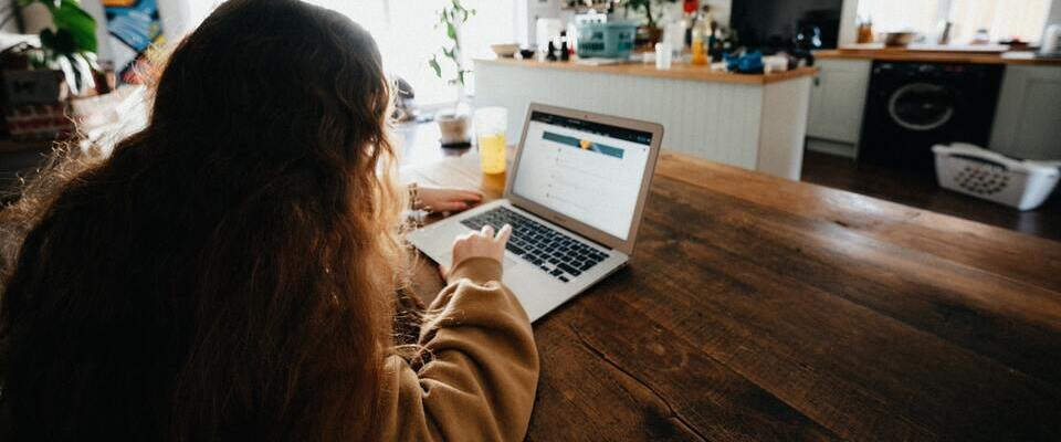 Pessoa fazendo revisão de texto na cozinha. O computador está em cima de uma mesa de madeira. 