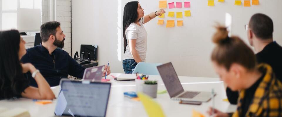 Pessoas da gestão de marketing fazendo uma reunião. Uma mulher está colando adesivos na parede enquanto seus colegas estão sentados em volta da mesa. 