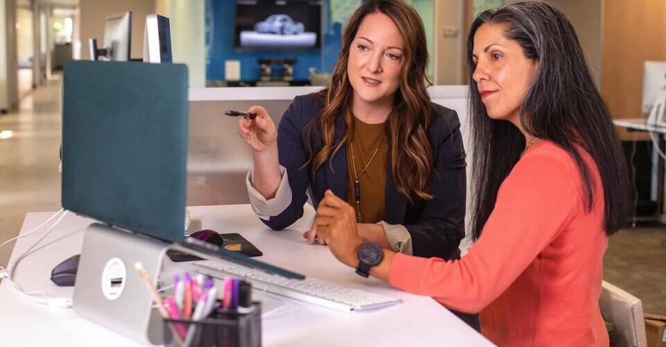 Duas mulheres na mesa olhando para tela do computador. Uma delas aponta com uma caneta para a tela. 