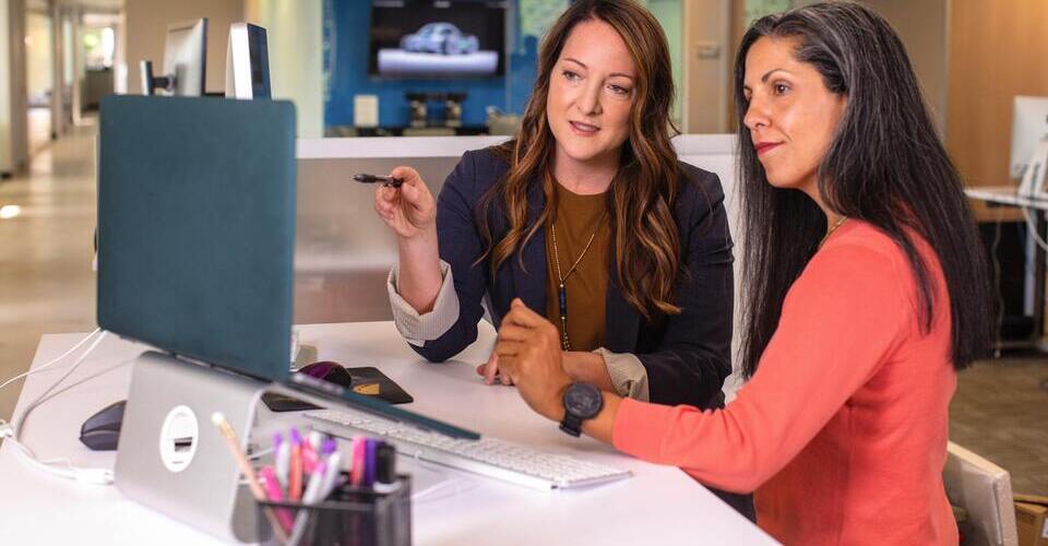 Duas mulheres na mesa olhando para tela do computador. Uma delas aponta com uma caneta para a tela.