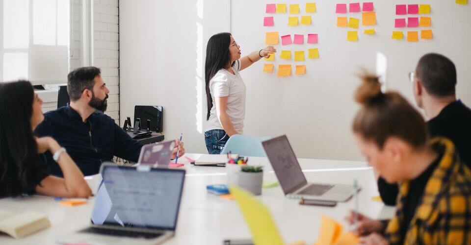 Reunião com várias pessoas ao redor de uma mesa branca. Na parede, vários post-its colados. 