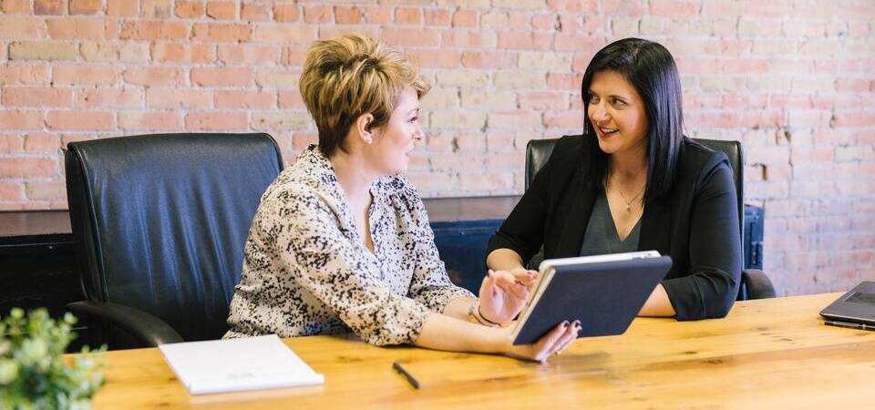Duas mulheres em uma mesa de reunião conversando e usando um tablet.