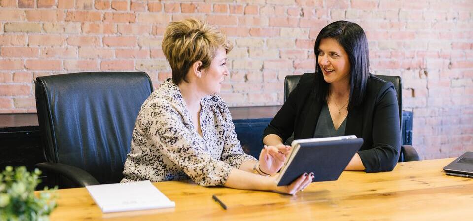 Duas mulheres em uma mesa de reunião conversando e usando um tablet. 