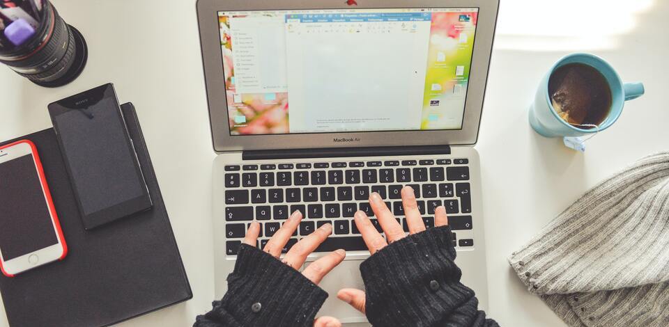 Pessoa digitando em um notebook. Na mesa há ainda celulares, caneca com chá, canetas e um gorro.