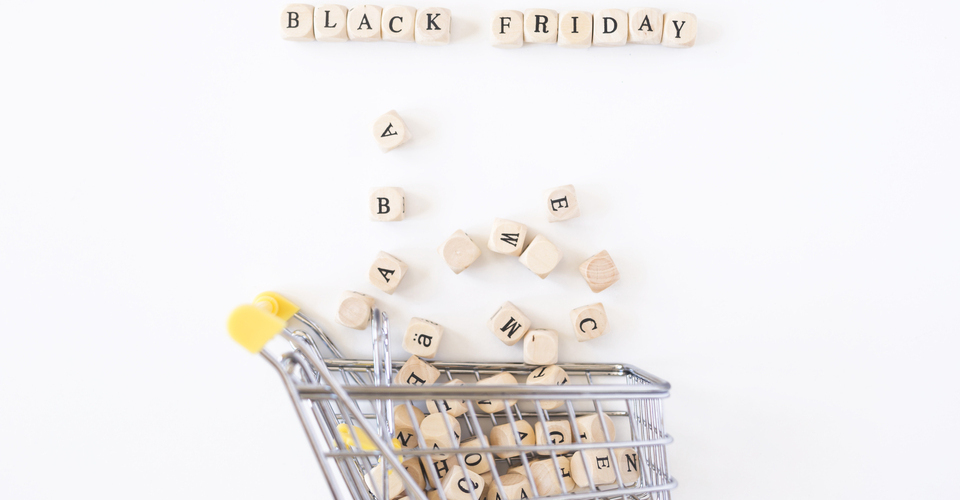 Imagem conceitual de um carrinho de compras em miniatura com vários blocos com letras espalhados por ele. Acima, a palavra “black friday”. 