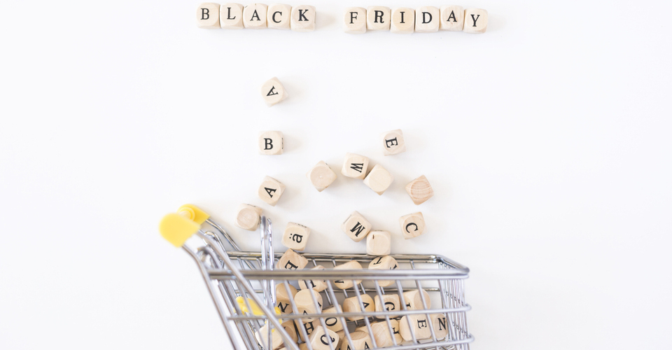 Imagem conceitual de um carrinho de compras em miniatura com vários blocos com letras espalhados por ele. Acima, a palavra “black friday”.