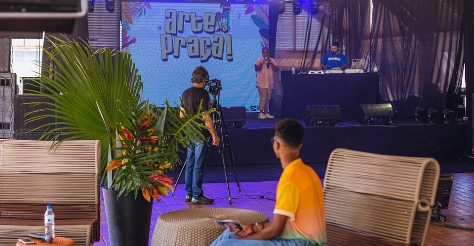 Apresentador sentado em poltrona da entrevista e operador de câmera filmando o show de Gehovanna Luiza, no palco do Arte na Praça. 