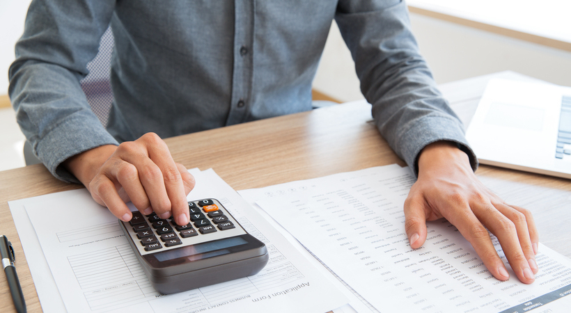 Foto de uma mão fazendo contas sem calculadora enquanto outra passa os dedos por lista de ítens a serem calculados.