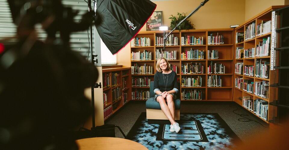 Foto de estudio de gravação montado na biblioteca de uma sala. No centro há uma mulher sentada em uma poltrona. 