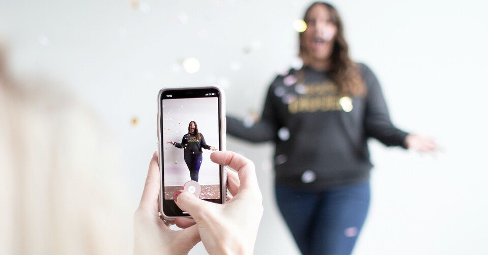 Foto de uma pessoa segurando smartphone. Na tela há uma mulher jogando confetes em si mesma.