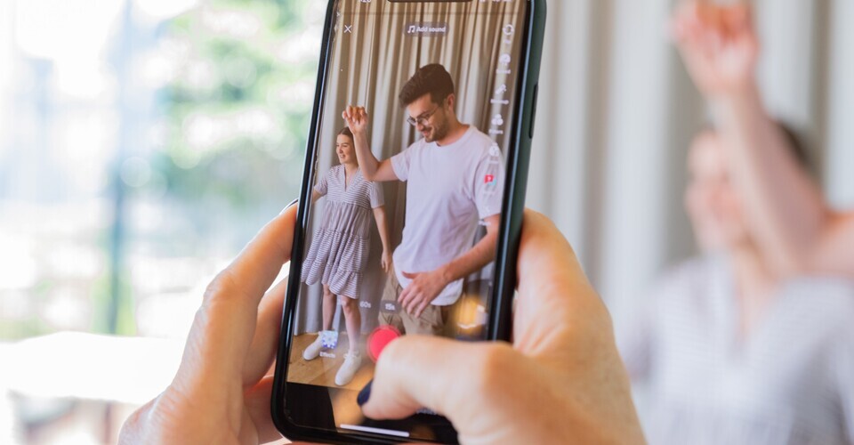 Foto de uma pessoa segurando smartphone. Na tela há duas pessoas fazendo danças descoordenadas. 