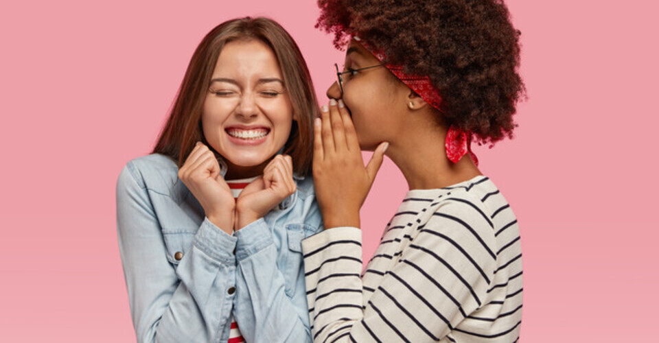 Duas garotas, uma de perfil contando algum segredo no ouvido de outra, que está de frente e animada com o que ouve. 