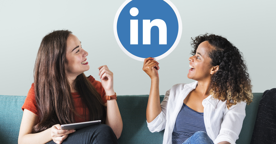 Foto de duas mulheres sorridentes sentadas no sofá. Uma está segurando placa com logotipo do LinkedIn e outra com um tablet na mão. 