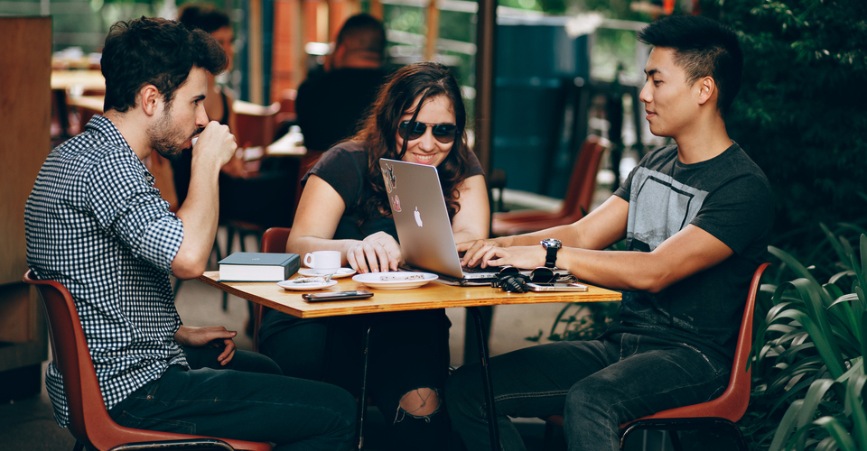 Foto de três pessoas em uma mesa ao ar livre. Eles estão tomando café, conversando e utilizando o notebook.