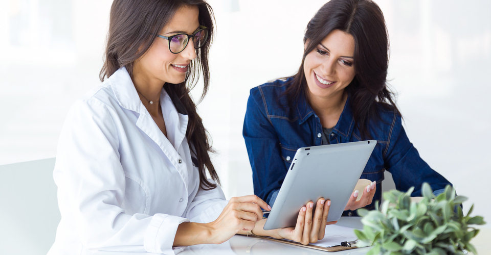 Duas mulheres sentadas analisando resultados em um tablet. Uma delas é profissional da saúde e usa jaleco.
