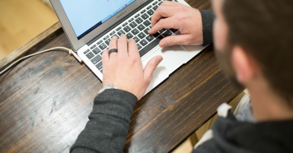 Foto de perspectiva superior com homem digitando em notebook que está em uma mesa de madeira.