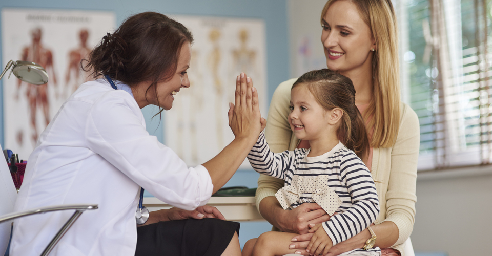 Médica, no consultório, fazendo saudação com criança que está sentada no colo da mãe.