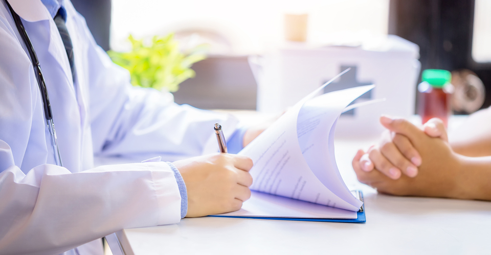 Médico assinando papéis sobre a mesa do consultório durante atendimento a paciente.