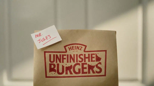 Sacola de papel Kraft com a logo da campanha “Unfinished Burgers” impresso em tinta vermelha. Há um post-it com “Mr. Jules” escrito. 