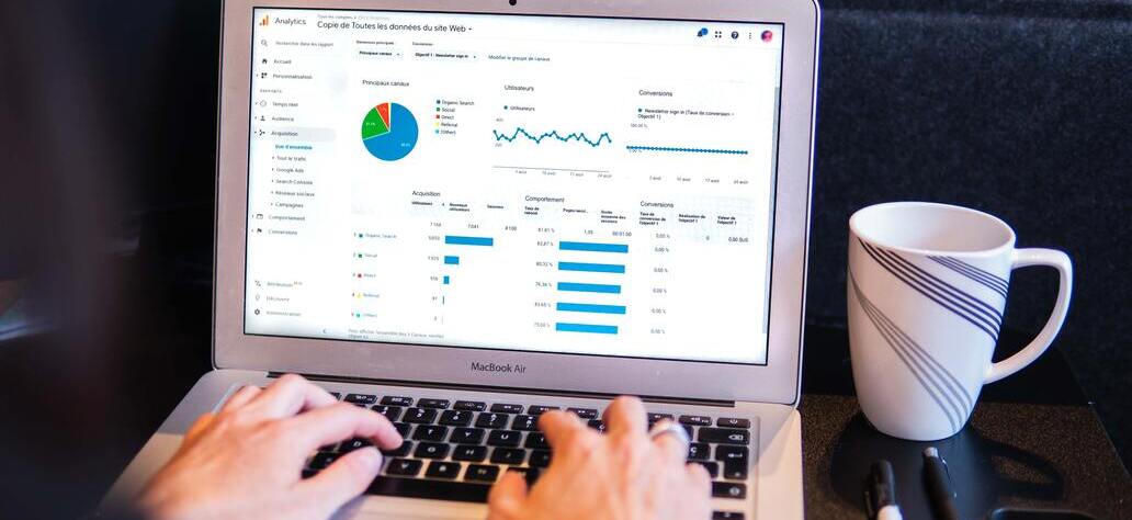 Pessoa usando o Google Analytics em MacBook Air. Ao lado, canetas e uma caneca.