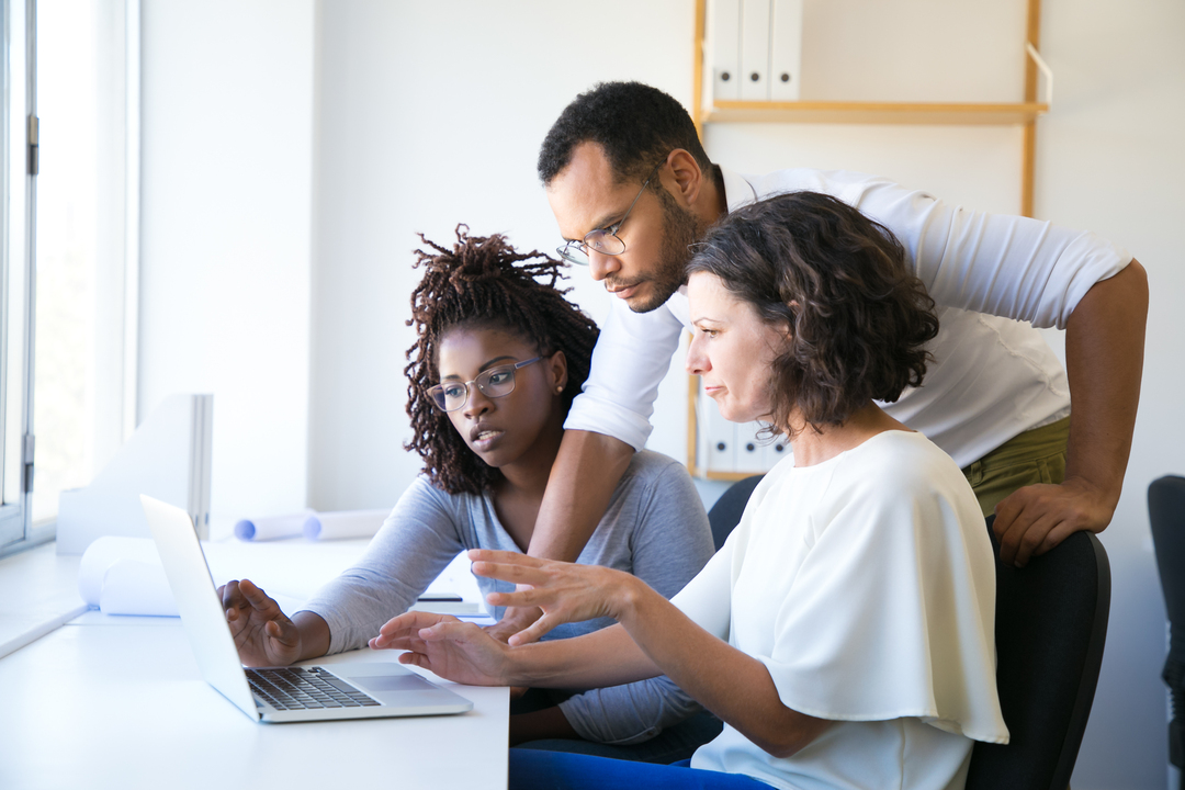 Foto de homem de negócios em pé ensinando duas mulheres com expressão confusa algo que está no notebook.