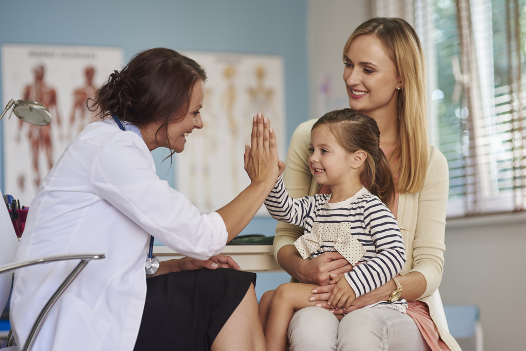 Médica, no consultório, fazendo saudação com criança que está sentada no colo da mãe.